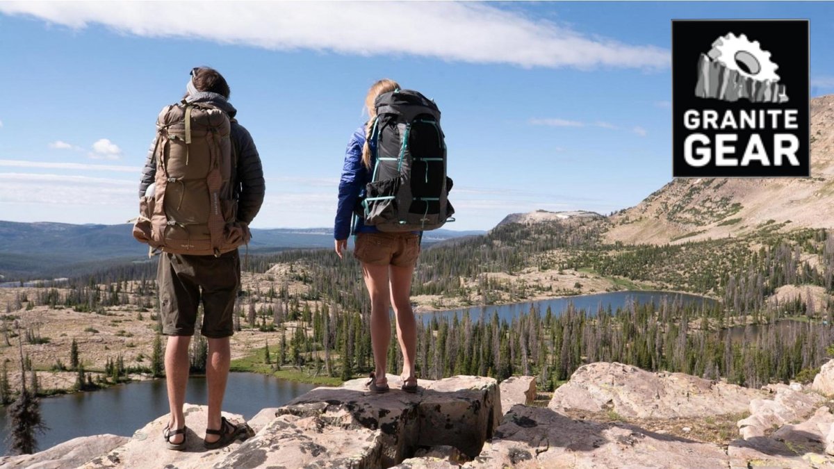 Two hikers wearing Granite backpacks stand on a rocky mountain overlook with alpine lakes and coniferous forest landscape below on a clear day.