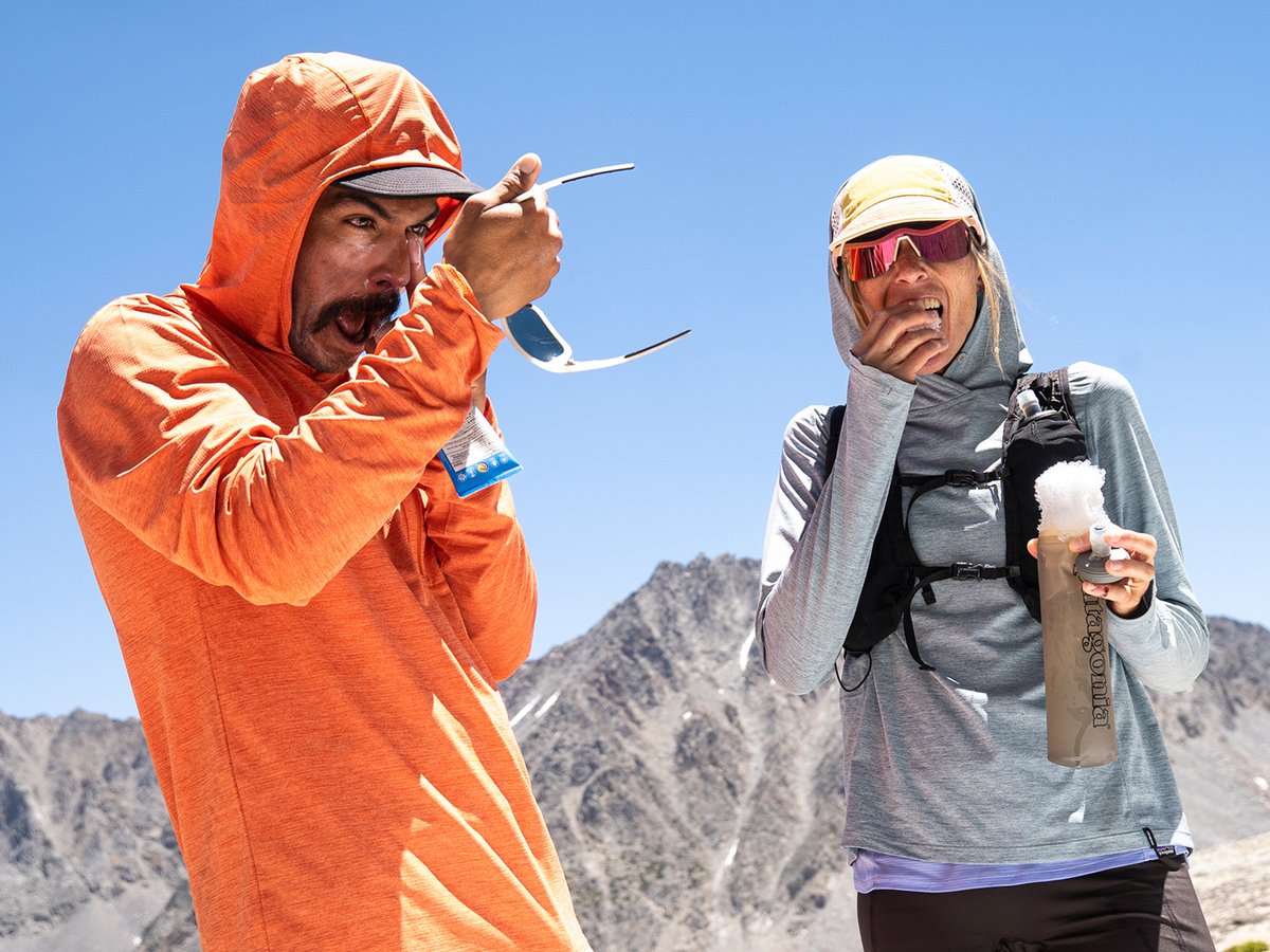Two mountain runners wearing brightly colored sun hoodies pause to snack and reapply sunscreen.