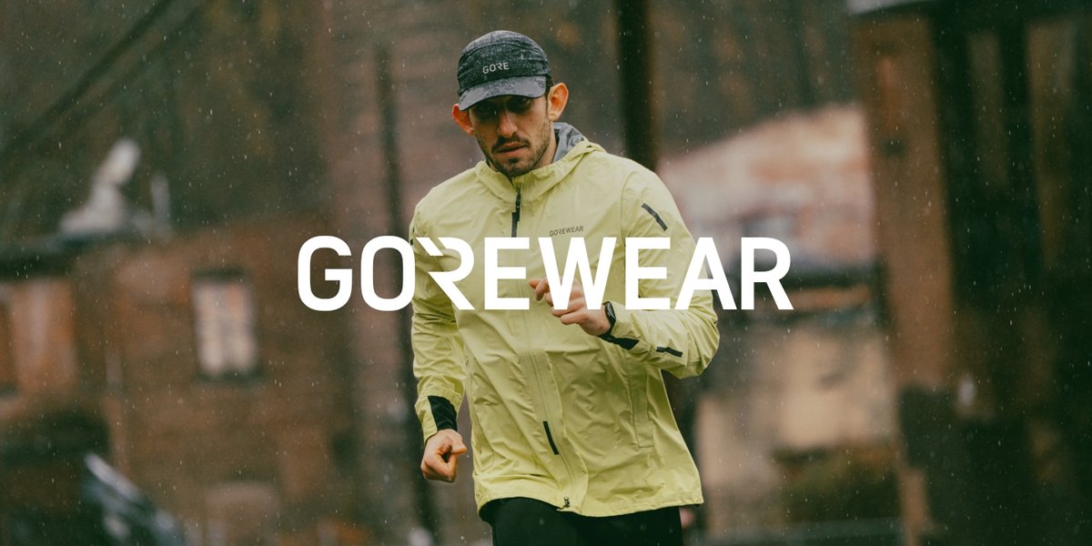 A person in a yellow GOREWEAR jacket and cap is running outdoors on a rainy day with buildings in the background.