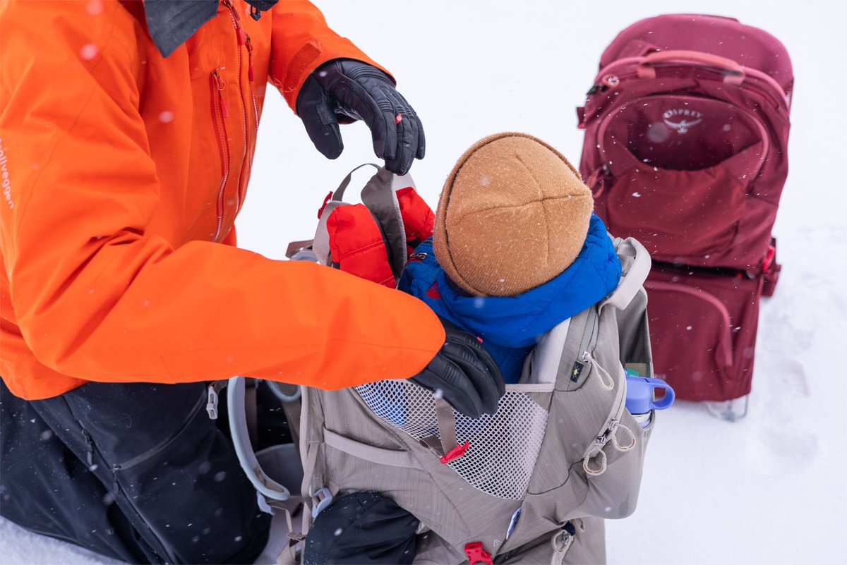 A person in an orange winter jacket and black gloves secures a toddler into an Osprey child carrier backpack in the snow. 