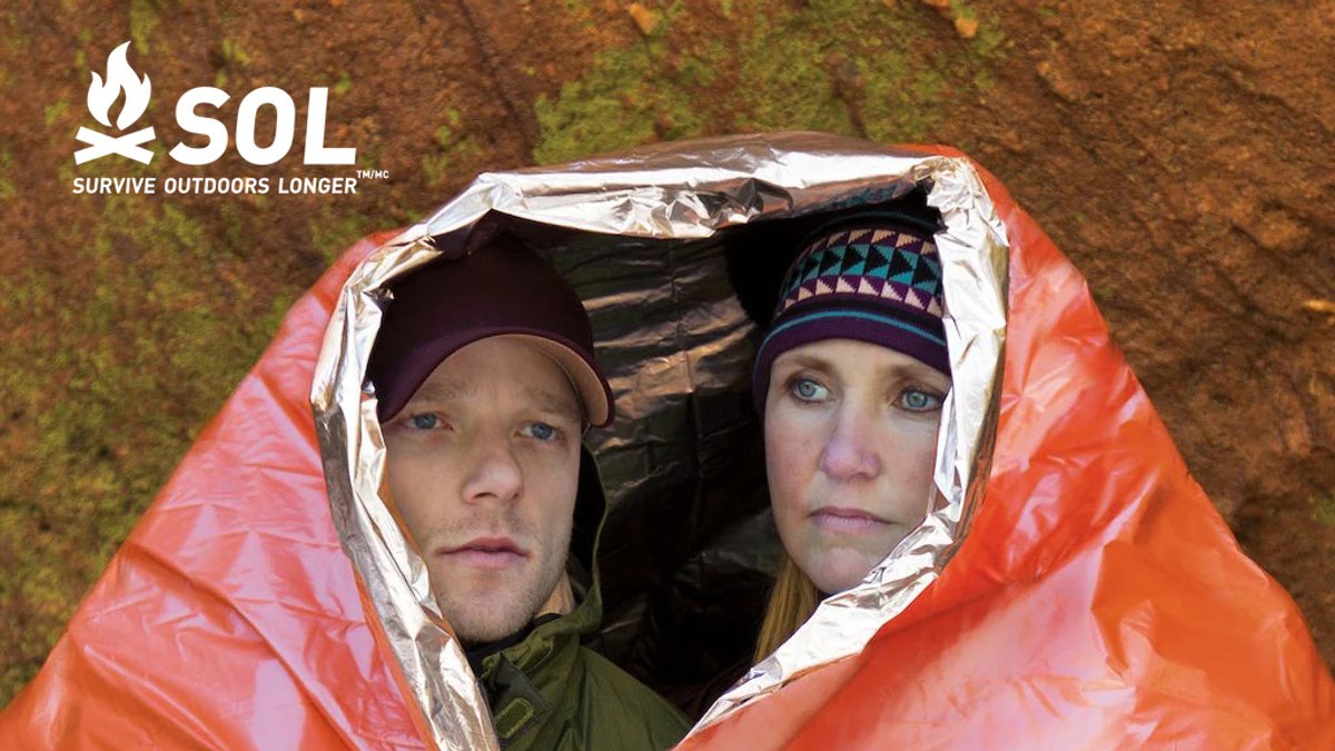 Two people huddled together inside a red and silver SOL emergency bivy shelter demonstrating warmth retention and compact emergency survival gear for outdoor protection.