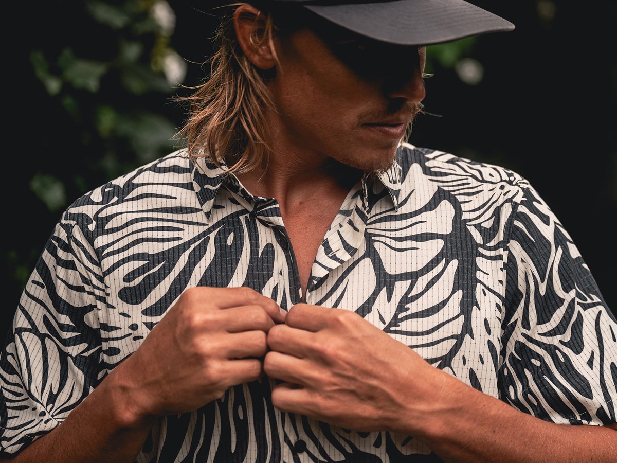 A man wearing a hat buttons up his shirt.