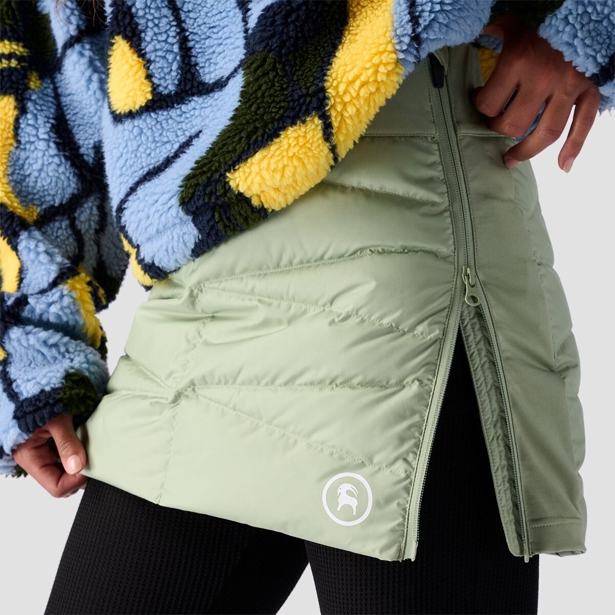Close-up of a woman wearing an olive Stansbury ALLIED Down jacket, showing the lofted quilted shoulder and hood with embroidered goat logo patch.