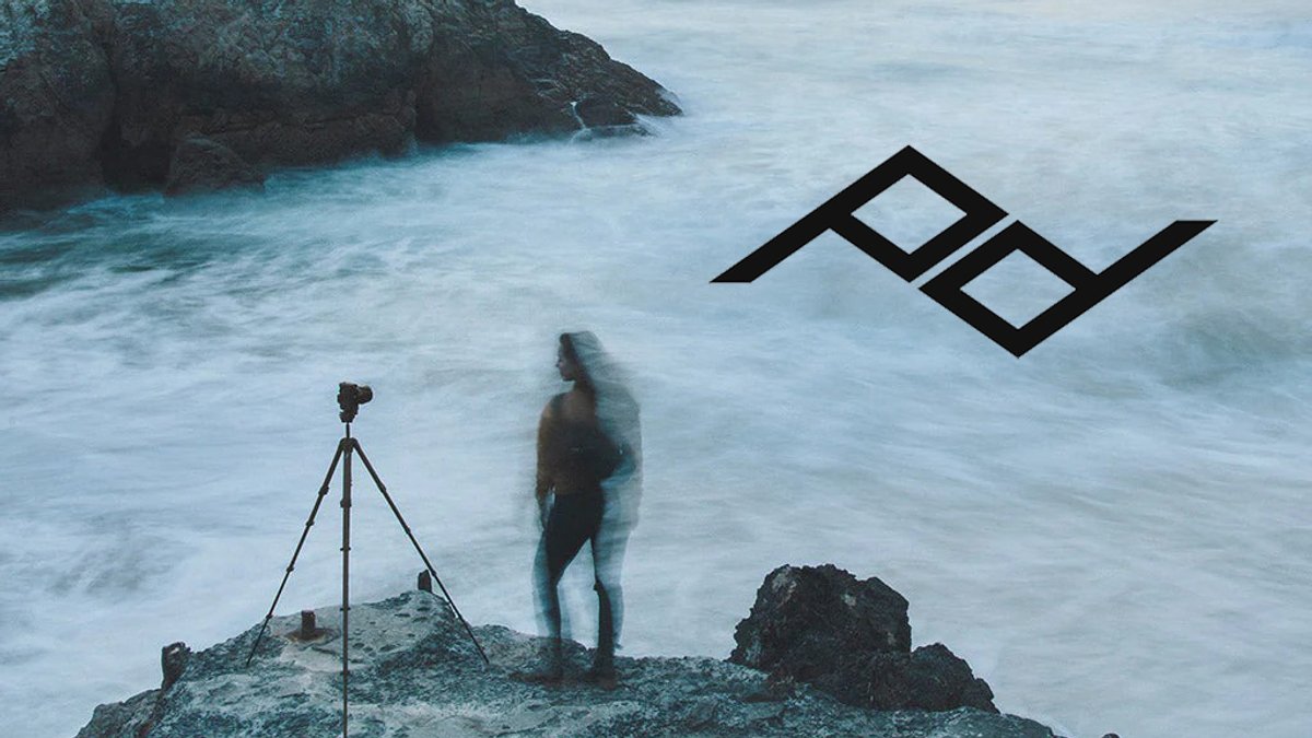 Long exposure coastal photography featuring a tripod-mounted camera capturing a wetsuit-clad surfer standing on rocky shoreline with crashing waves and dramatic cliffs in the background.