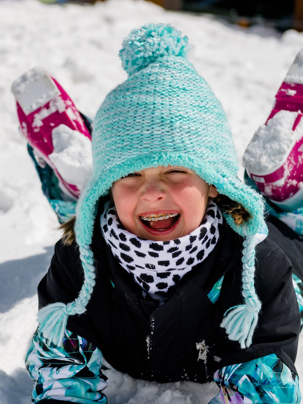 Sliding in the snow, a child wearing a light blue knit hat with a pom-pom and ear flaps, a black winter jacket, a black-and-white spotted neck gaiter.