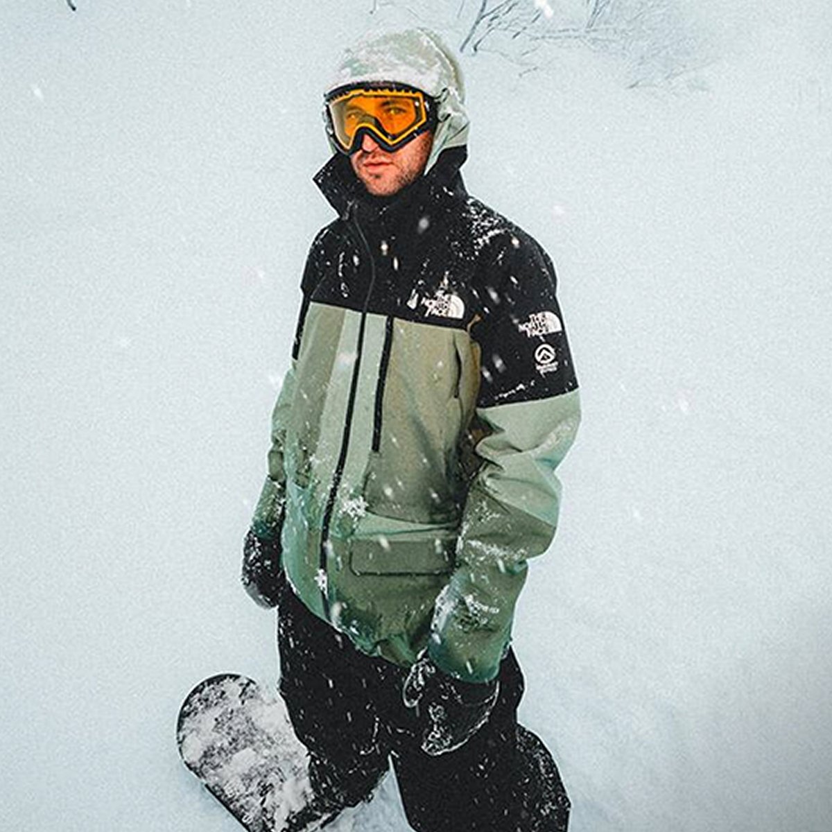 A person in a hooded winter jacket and yellow goggles stands on snow next to a snowboard.