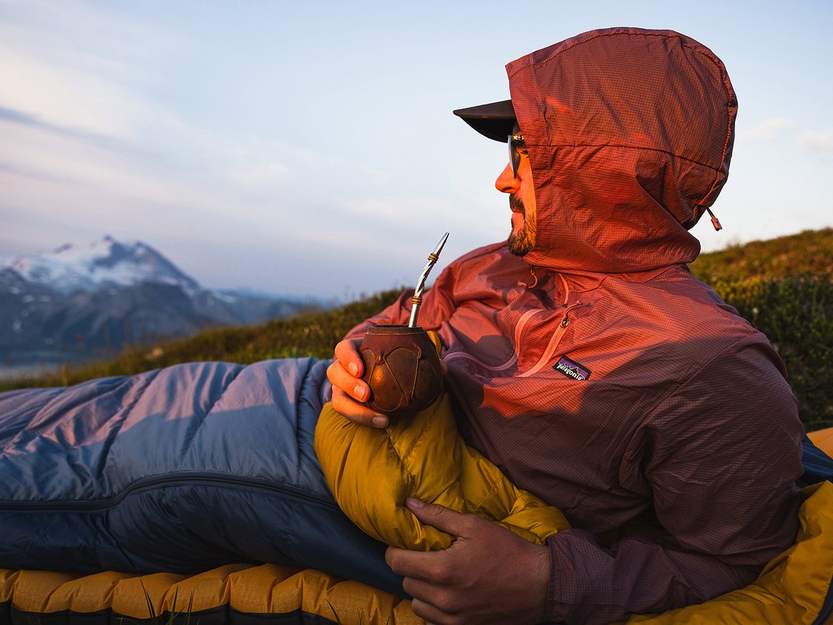 A man sips a drink while laying down in his sleeping bag.