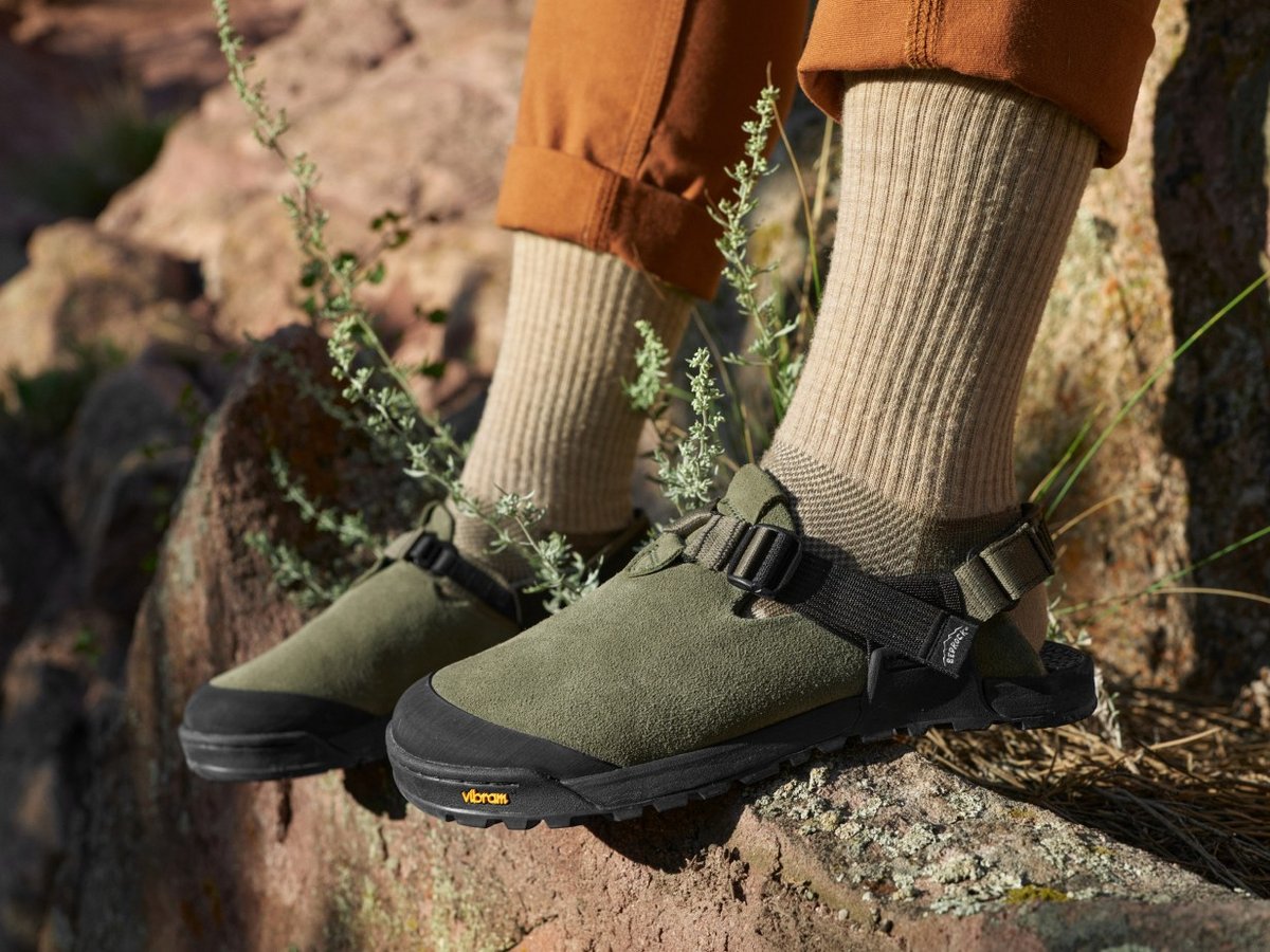 Closed-toe suede sandals with a black rubber toe cap and strap, worn with wool socks on a sunlit rock outcrop.