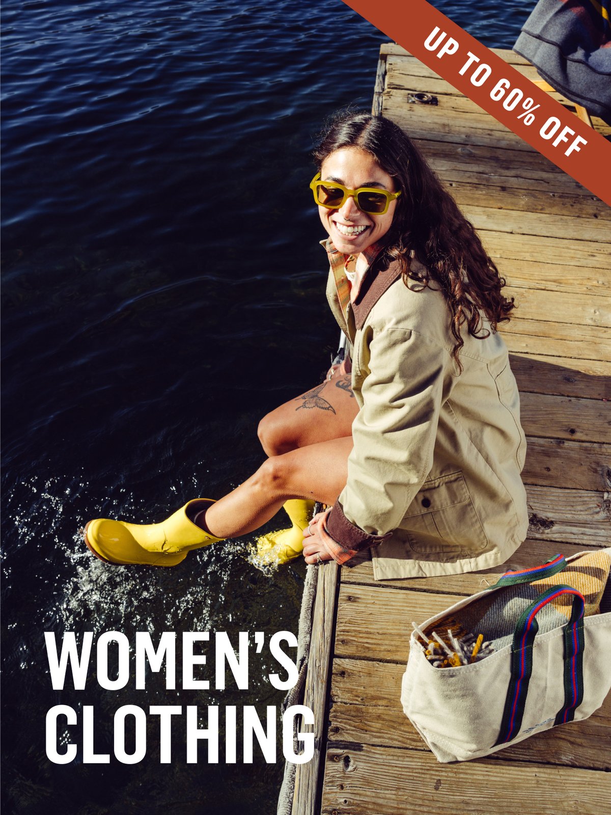 Woman sitting on a wooden dock, feet splashing in lake water, wearing a tan jacket, yellow rain boots, and yellow-green sunglasses. Women's Clothing Up To 60% off. 
