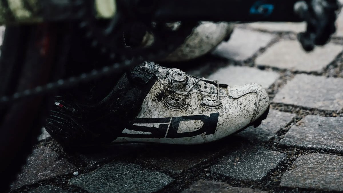 A Sidi cycling shoe is covered in dust and mud on a gravel road, showcasing durability and performance in off-road terrain.