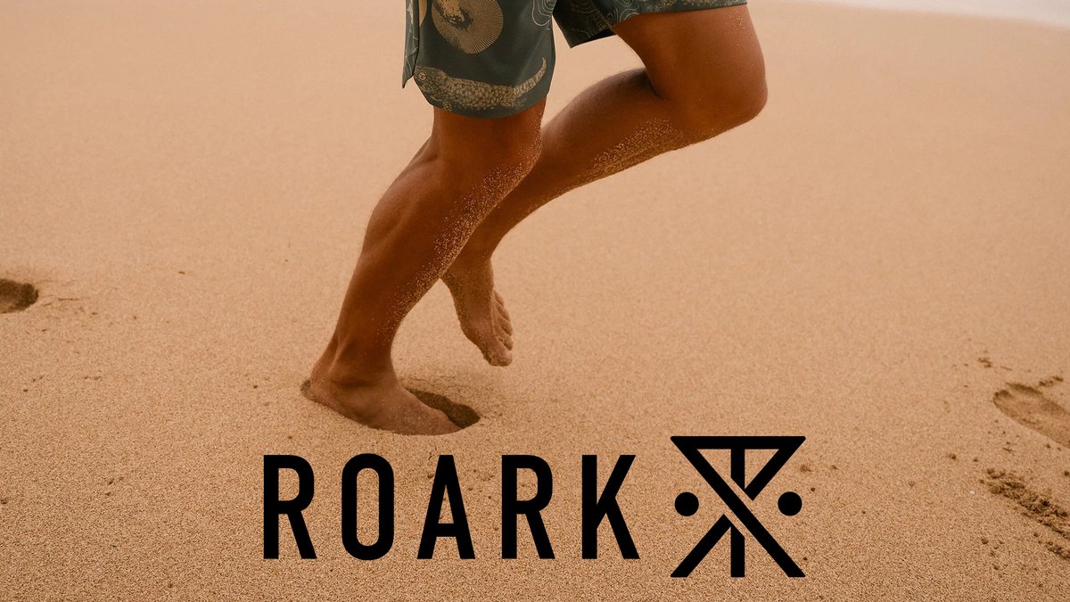 A close-up shot of a person's legs and bare feet running across a sandy beach with the Roark brand logo at the bottom.
