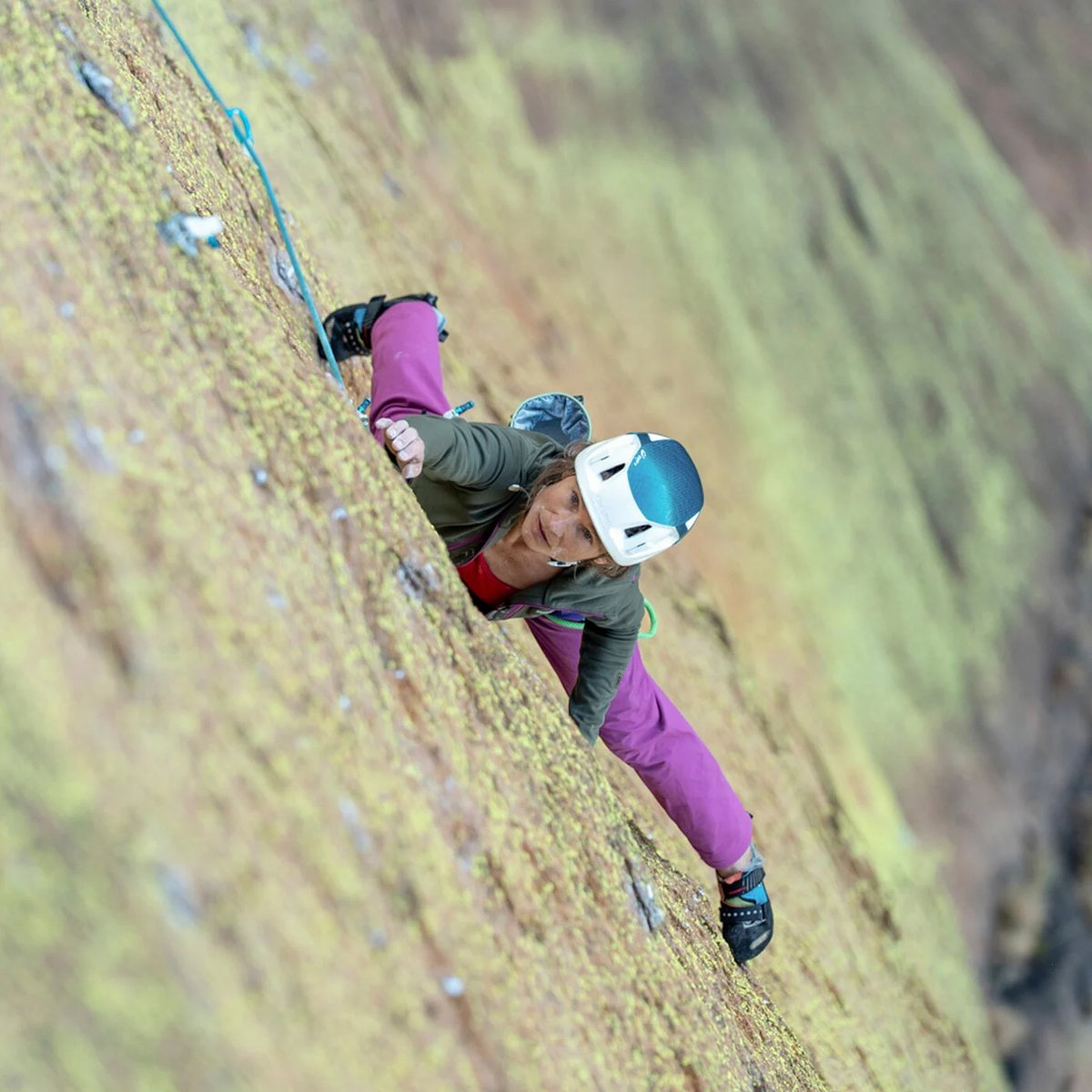 A woman climbs up a cliff face, looking up as she thinks intensely about her next move.