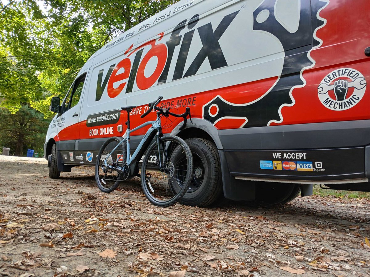 A velofix van is on a forest road with fallen leaves, while a bike leans against the van.