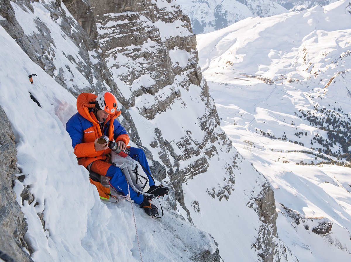 A mountaineer wearing bright orange and blue Mammut outerwear rests between pitches on a snowy cliff face.