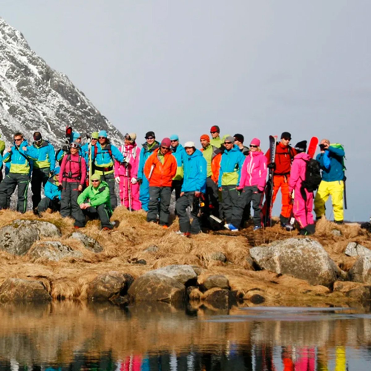 A large group of tourists wearing colorful waterproof jackets and winter gear stands together on a rocky mountain slope overlooking a calm lake during an Arctic hiking excursion.