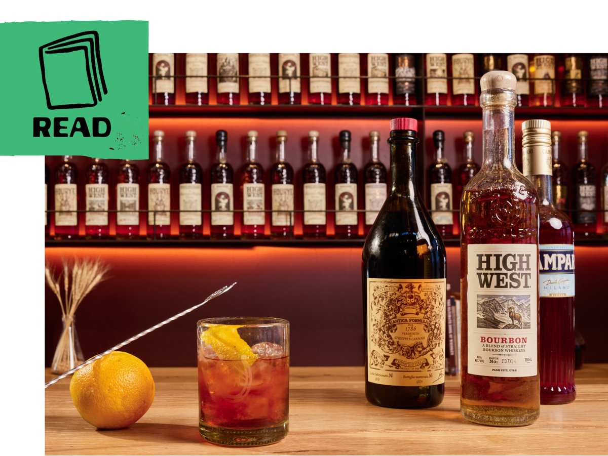 A bar countertop displays a red cocktail in a rocks glass beside a bottle of High West Bourbon, a bottle of sweet vermouth, and a bottle of Campari. A green flag includes an illustration of a book and the word “read.”
