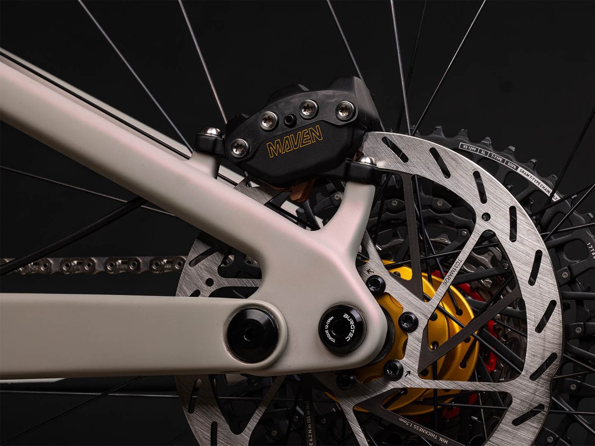 Rear disc brake caliper detail labeled "Maven," mounted near a gold hub and cassette, with rotor and spokes visible.