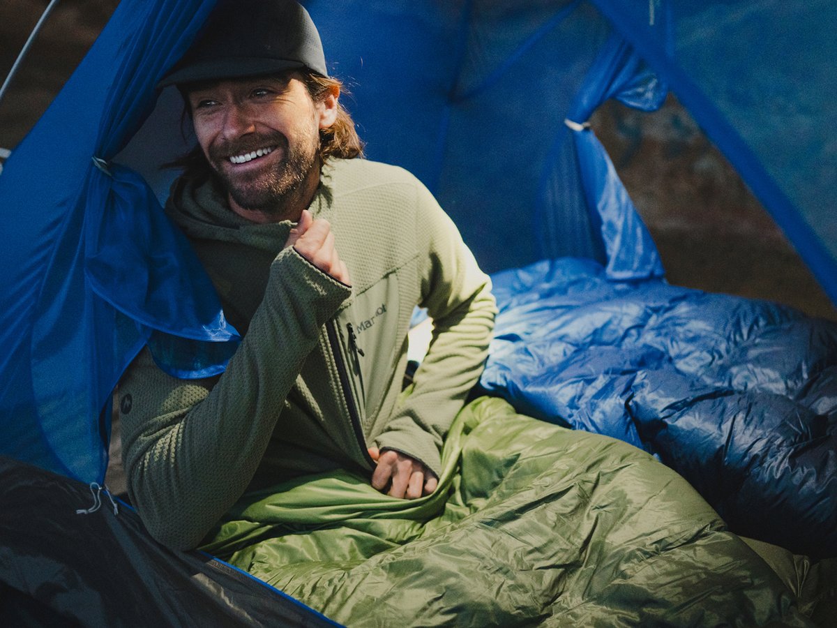 A man settling into his green sleeping bag inside a two-person tent.