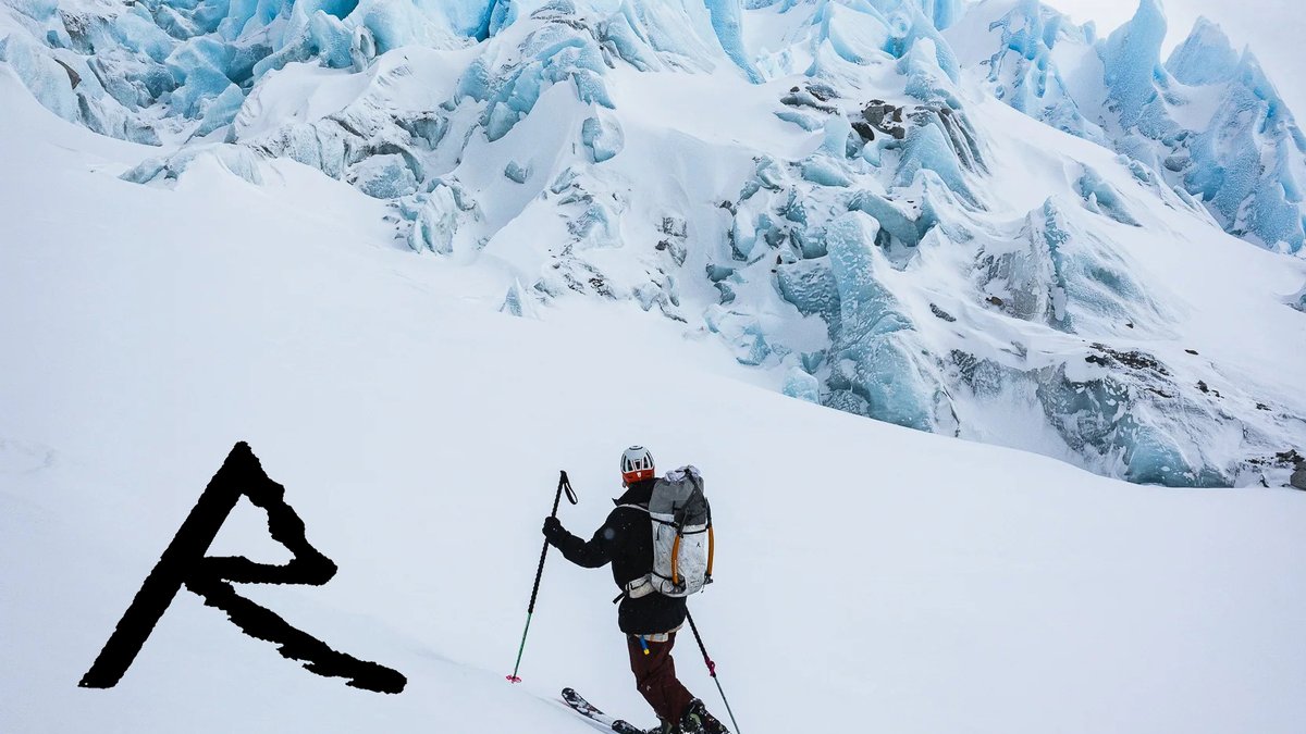 A skier ascends a snowy slope with a large crevassed glacier face looming in the background.
