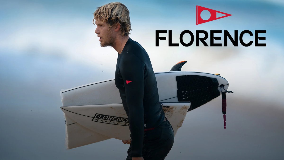 Male surfer in black wetsuit carrying a Florence Marine X surfboard with red flag logo on misty beach.
