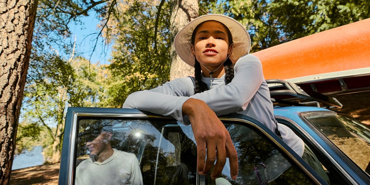 A woman wearing a sun shirt and large-brimmed hat rests against a car.