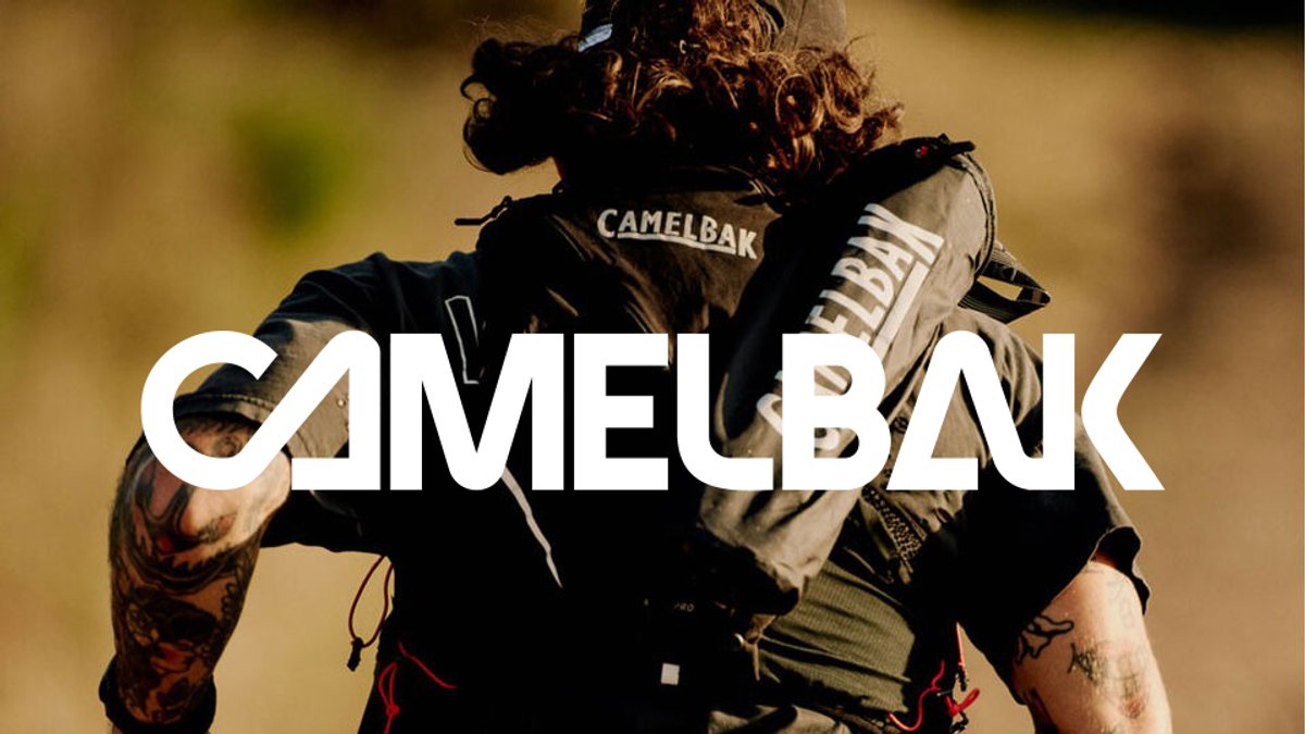 A person wearing a CamelBak hydration backpack and tactical gear stands outdoors with a blurred natural background.
