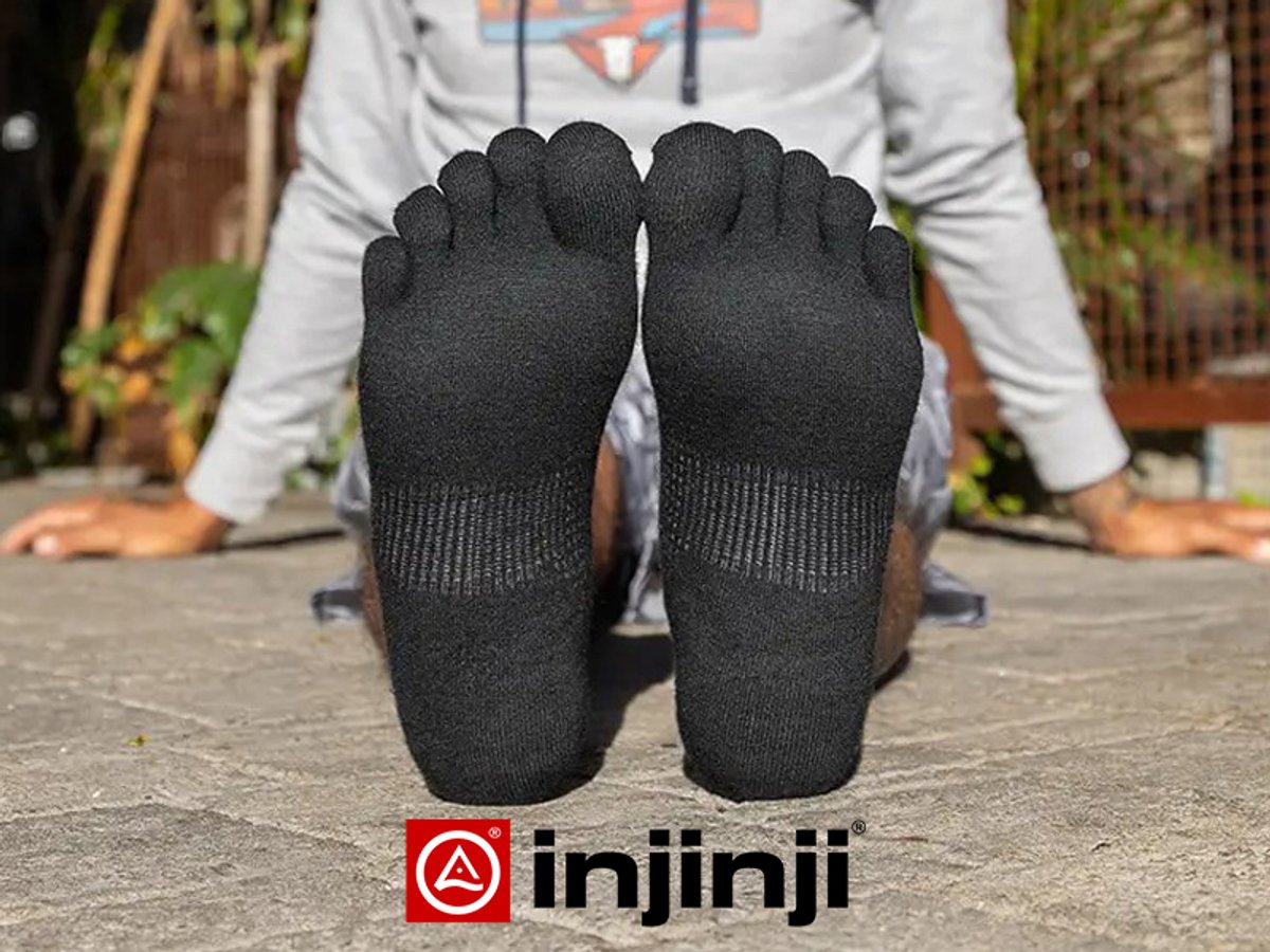 A close-up view of the soles of black Injinji toe socks being worn by a person sitting outdoors; the Injinji logo is at the bottom.