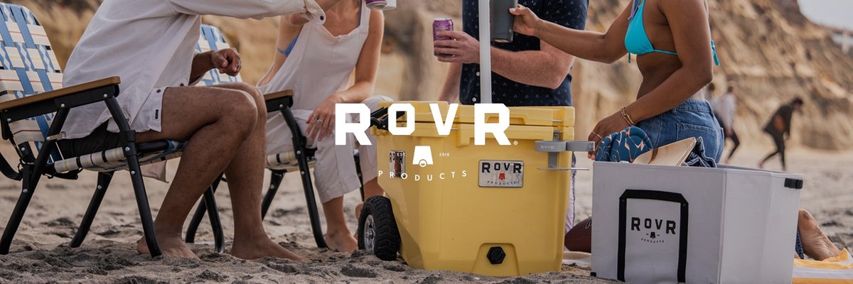 Three people sit and kneel on a sandy beach near rocky cliffs, relaxing with drinks and snacks next to two coolers. The ROVR logo is overlaid. 