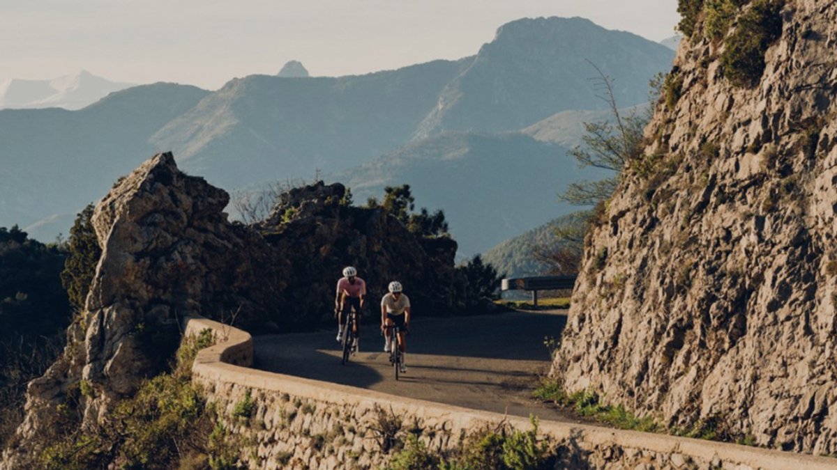 Two cyclists ride along a narrow, winding mountain road carved into a steep rocky cliffside.