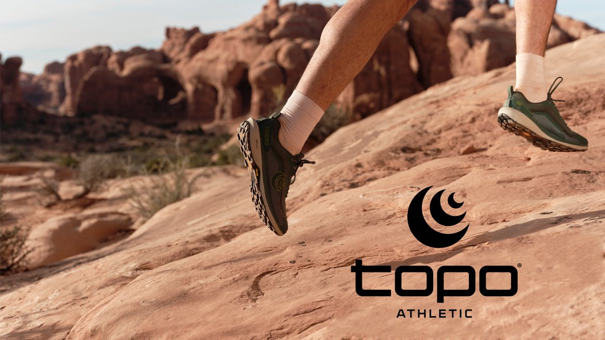 Runner wearing olive green Topo Athletic trail shoes with white socks jogging across red desert rock formations in a scenic outdoor landscape.