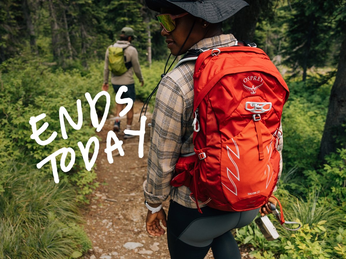 Two people wearing backpacks hike through the woods on a dirt path.