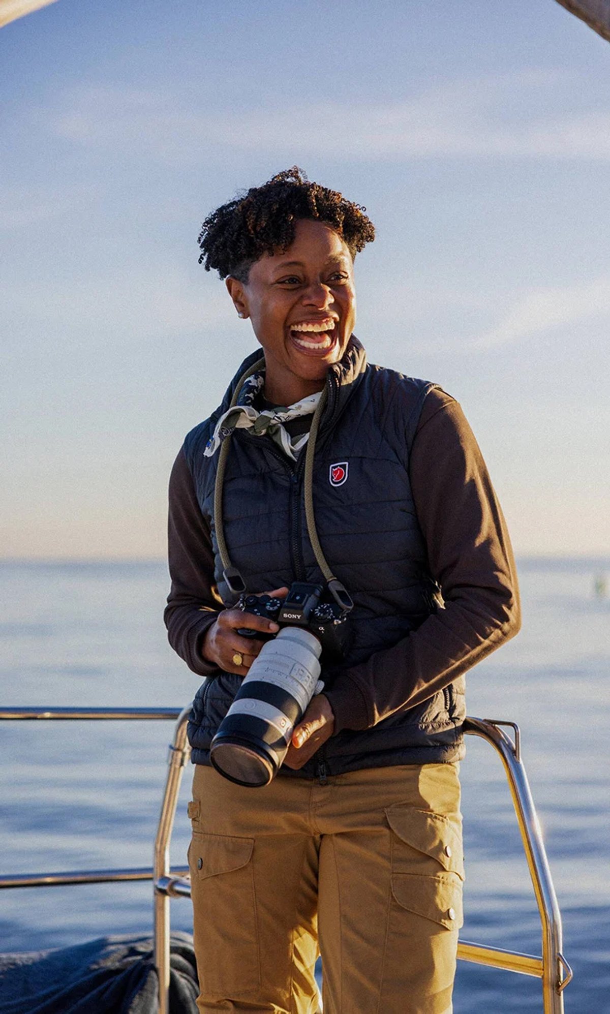 A woman wears the Fjallraven Expedition vest. She holds a camera while standing on a deck with a boat.
