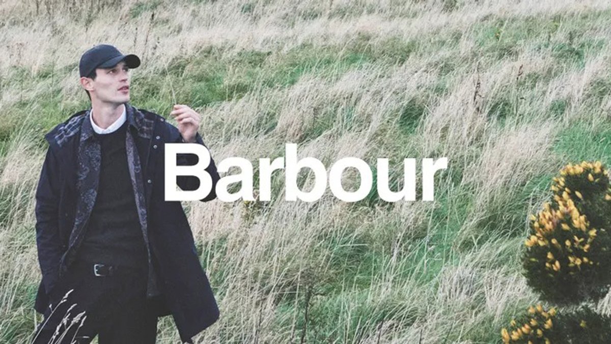 A man in a dark Barbour jacket and cap stands in a field of tall, windblown grass with the brand name centered in white.