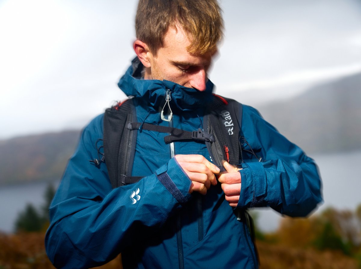 A man wearing a dark blue Rab rain jacket and a black backpack is outdoors, adjustss his jacket in a misty, mountainous landscape near a body of water.