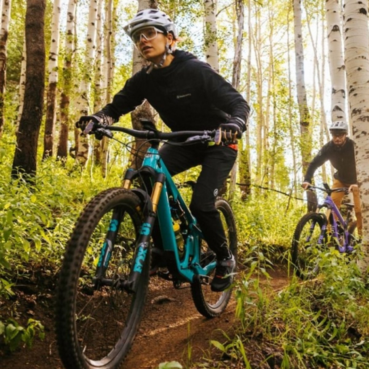 Two mountain bikers pedal along a wooded trail. 