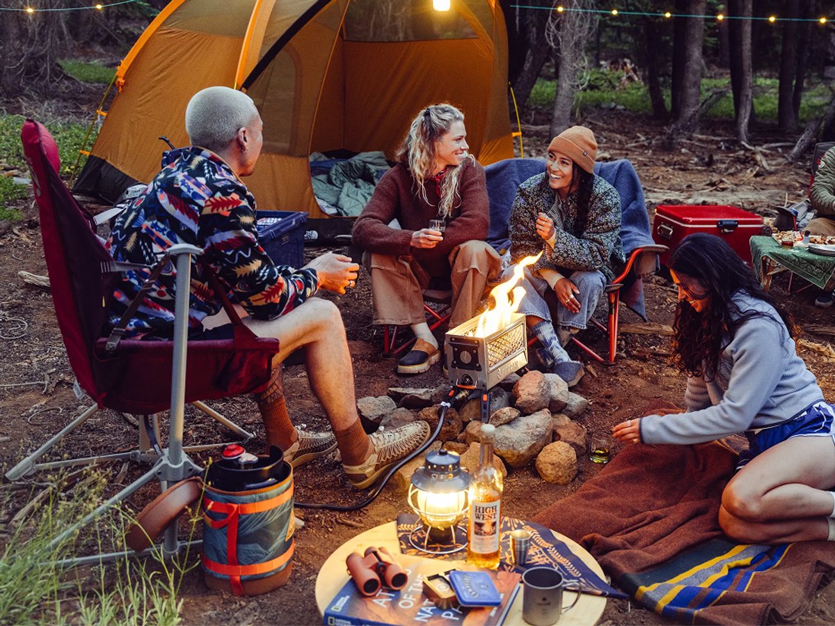 Five friends gathered around a small camp stove fire in a pine forest, laughing and sharing whiskey beside a glowing orange tent.