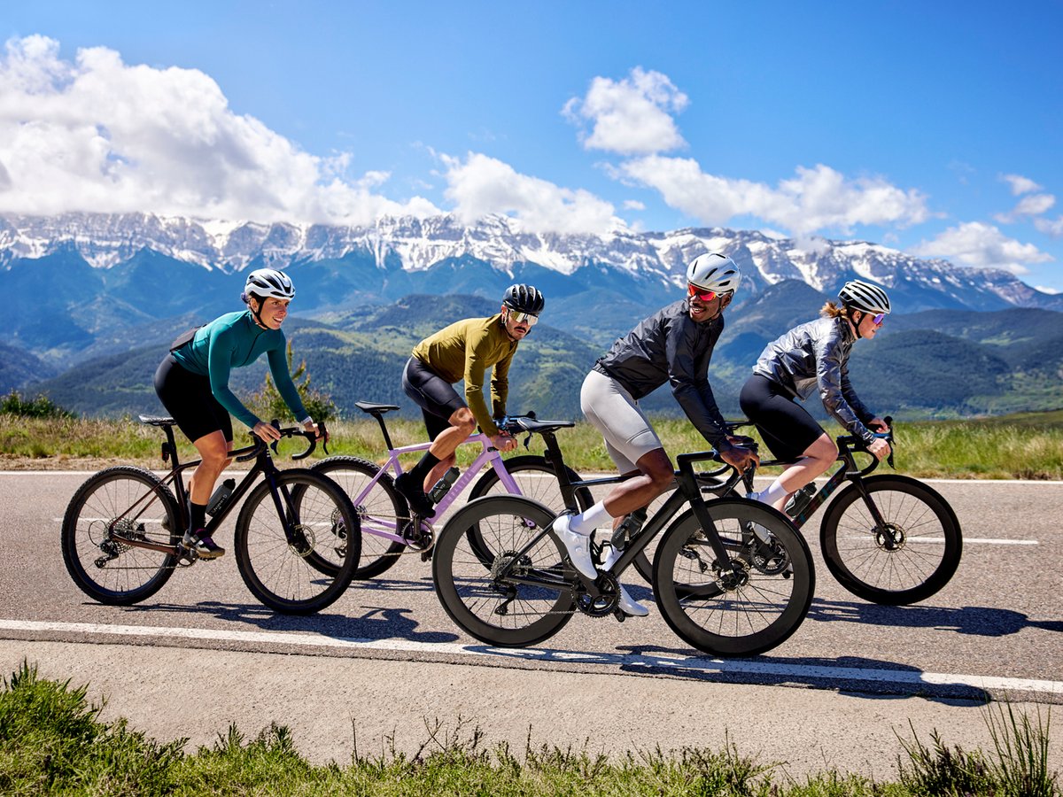 A group of four people on road bikes ride against a blue sky and snow-capped mountains.
