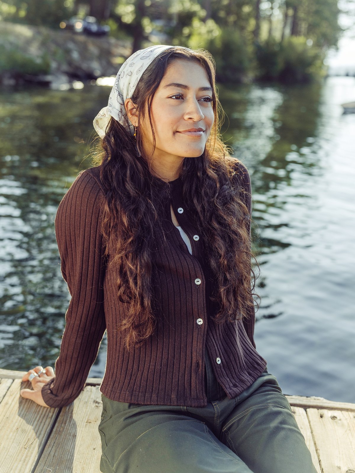 A woman with long wavy hair wearing a white bandana, a ribbed dark brown button-up cardigan, and olive green pants, sits on a wooden dock by the water with trees in the background.