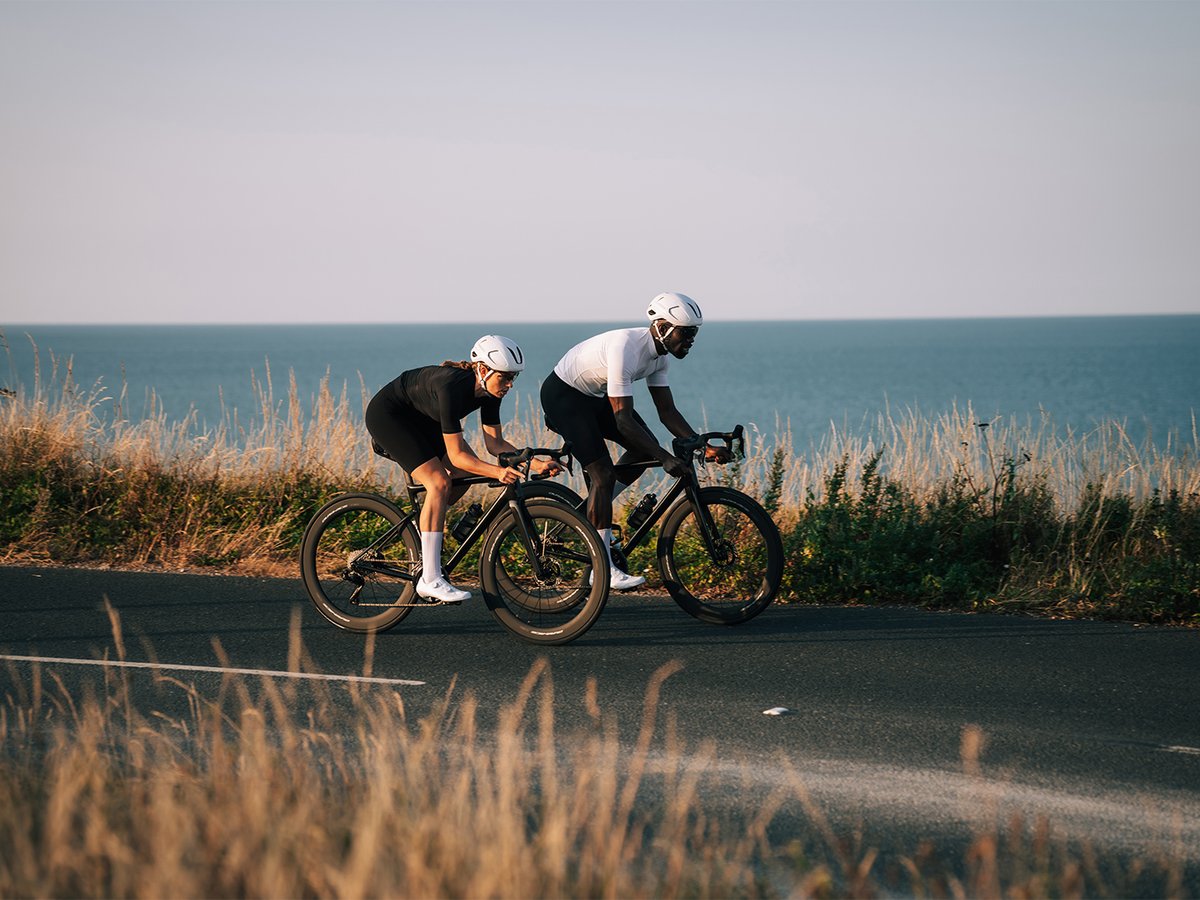 Cyclist in black kit riding along a coastal road, Dura-Ace wheels visible, white S-PHYRE shoe and sock visible, dry grass and ocean in the background