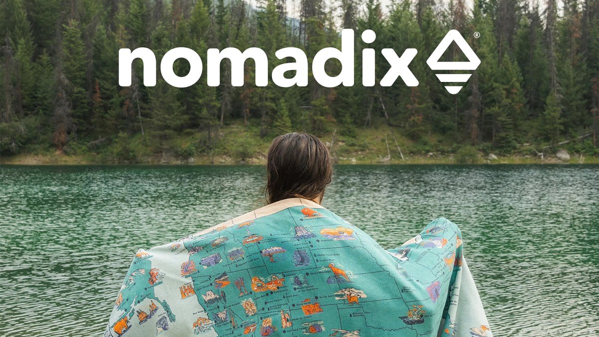 Person wrapped in a colorful Nomadix map towel overlooking a pristine alpine lake surrounded by evergreen forest.