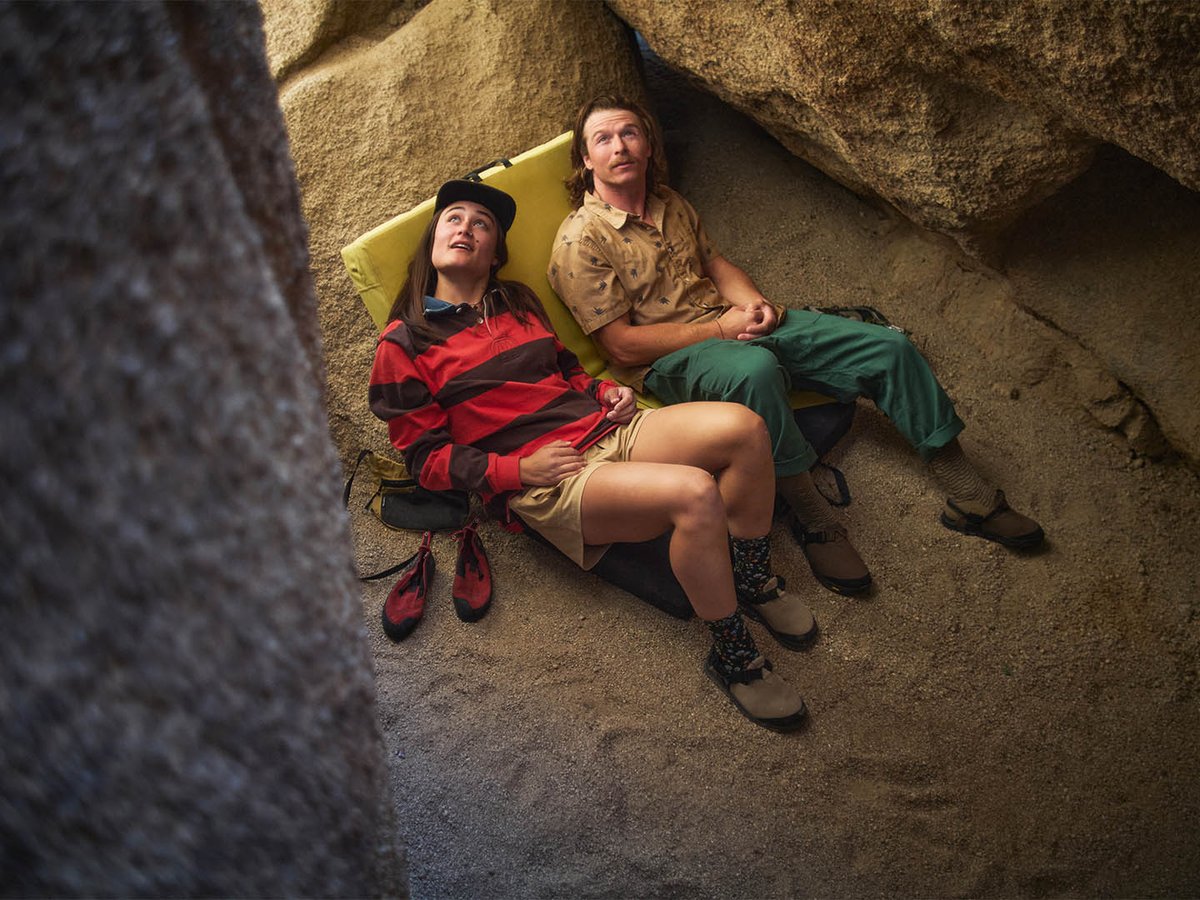 Two people resting on a sleeping pad beneath granite boulders wearing Bedrock Mountain Clogs, with a pair of red shoes set beside them