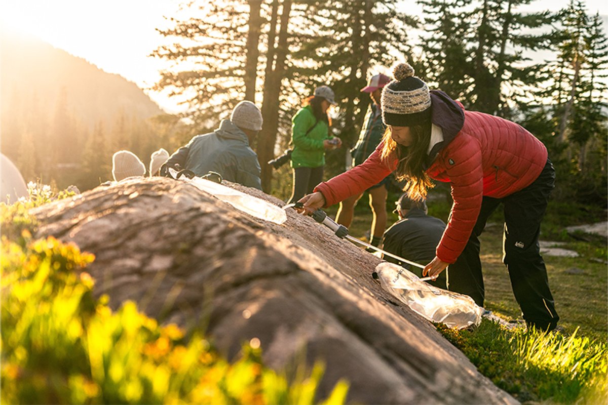 Camper sets up a gravity water filter on a rock at a forested campsite during golden-hour light, with others in the background.
