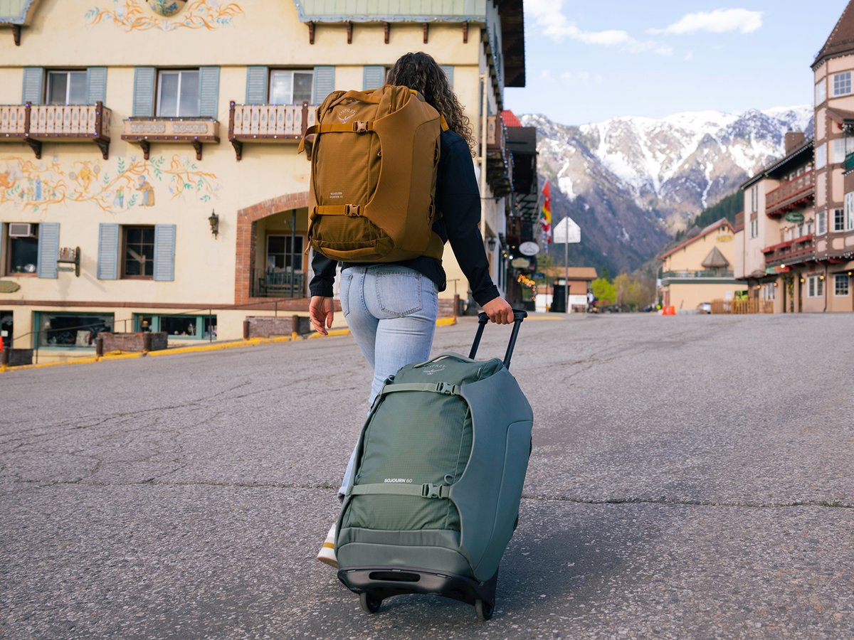 Woman walking through a mountain town street with a green rolling duffel and tan backpack, snow-capped peaks visible in the background.