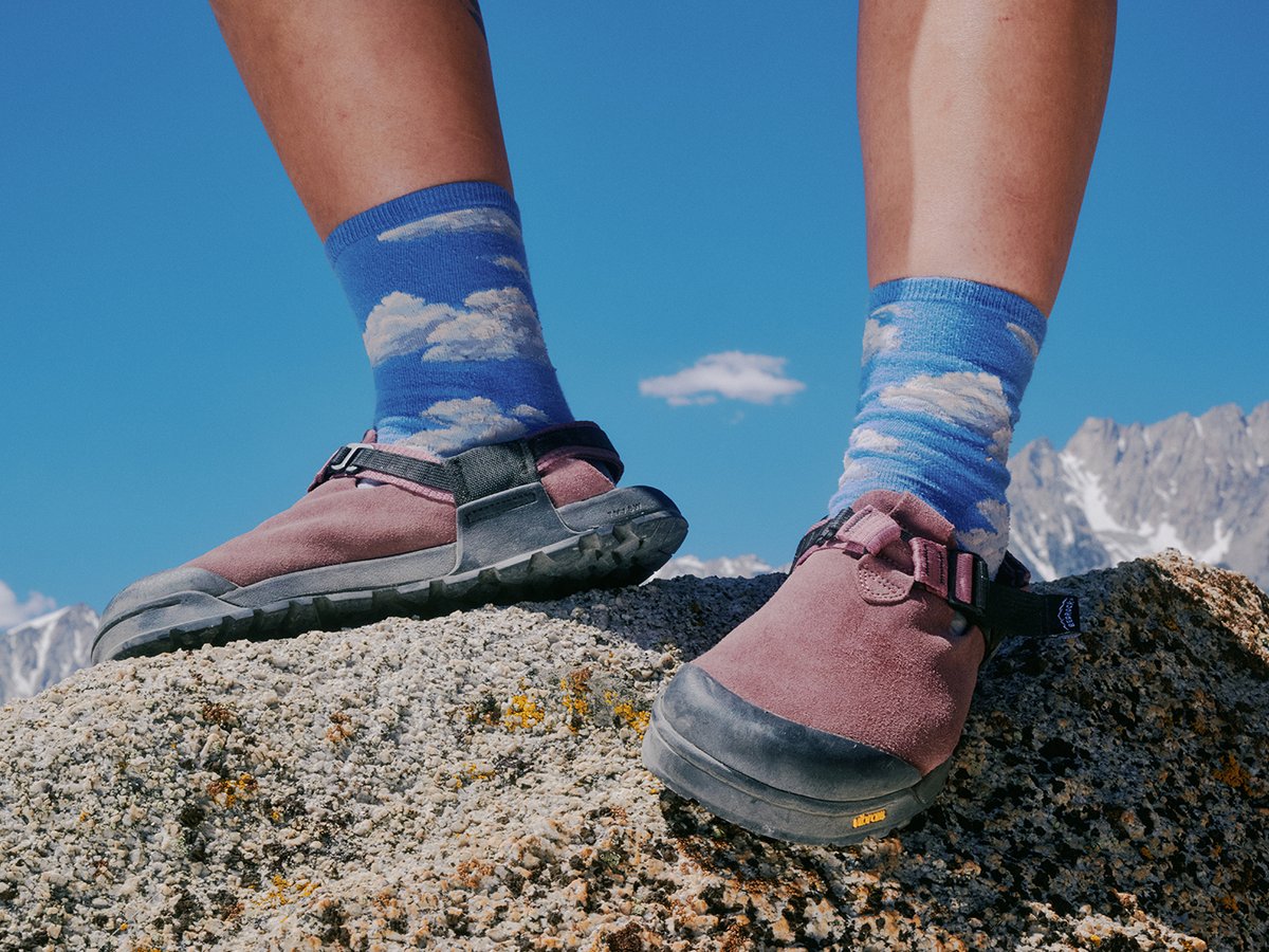 Low-angle view of Bedrock Mountain Clogs in pink with blue socks standing on rock, showing rubber toe cap