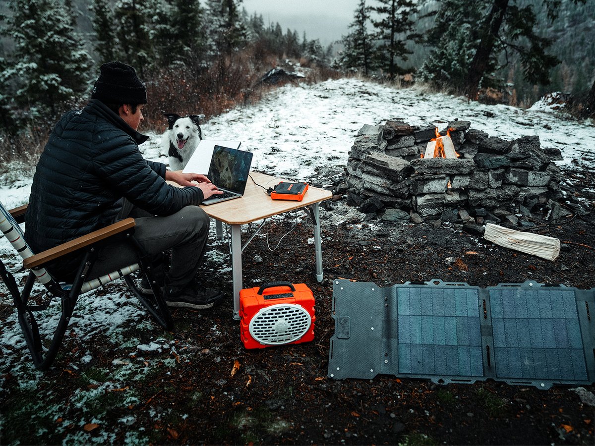 A camper works on a laptop at a folding table beside a stone fire ring in light snow, powered by an orange Dark Energy power station with a Spectre solar panel unfolded on the ground and a dog resting nearby.