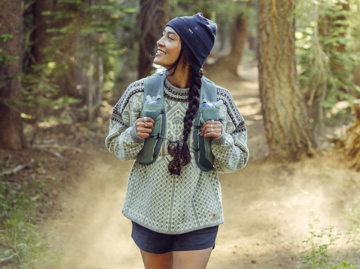 A person with a long braid and a navy beanie walks on a forest trail, wearing a patterned knit sweater and carrying a hiking vest with water bottles and sunglasses attached.