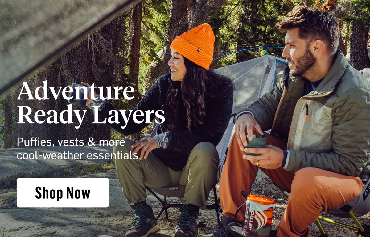 A woman and a man dressed in fleece jackets and vest share coffee at camp. Overlaid text reads, “Shop adventure ready layers, like puffies, vests & more cool-weather essentials”