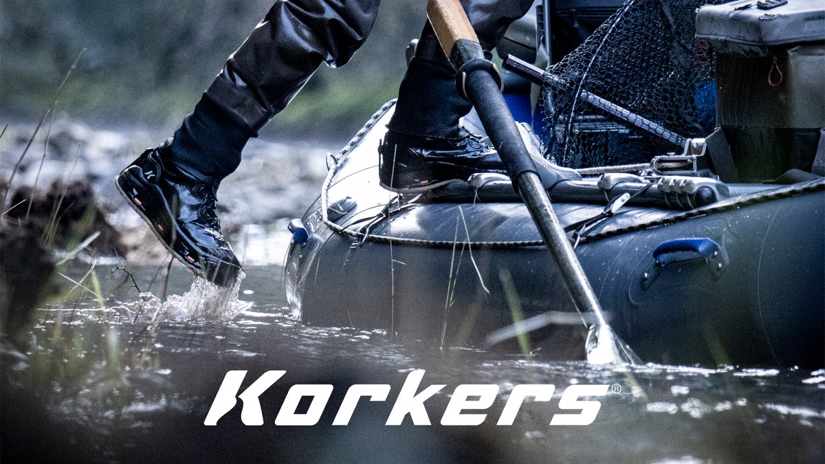 Person wearing waterproof Korkers boots wading through river water while fishing with net and gear, demonstrating durable outdoor footwear for water activities.