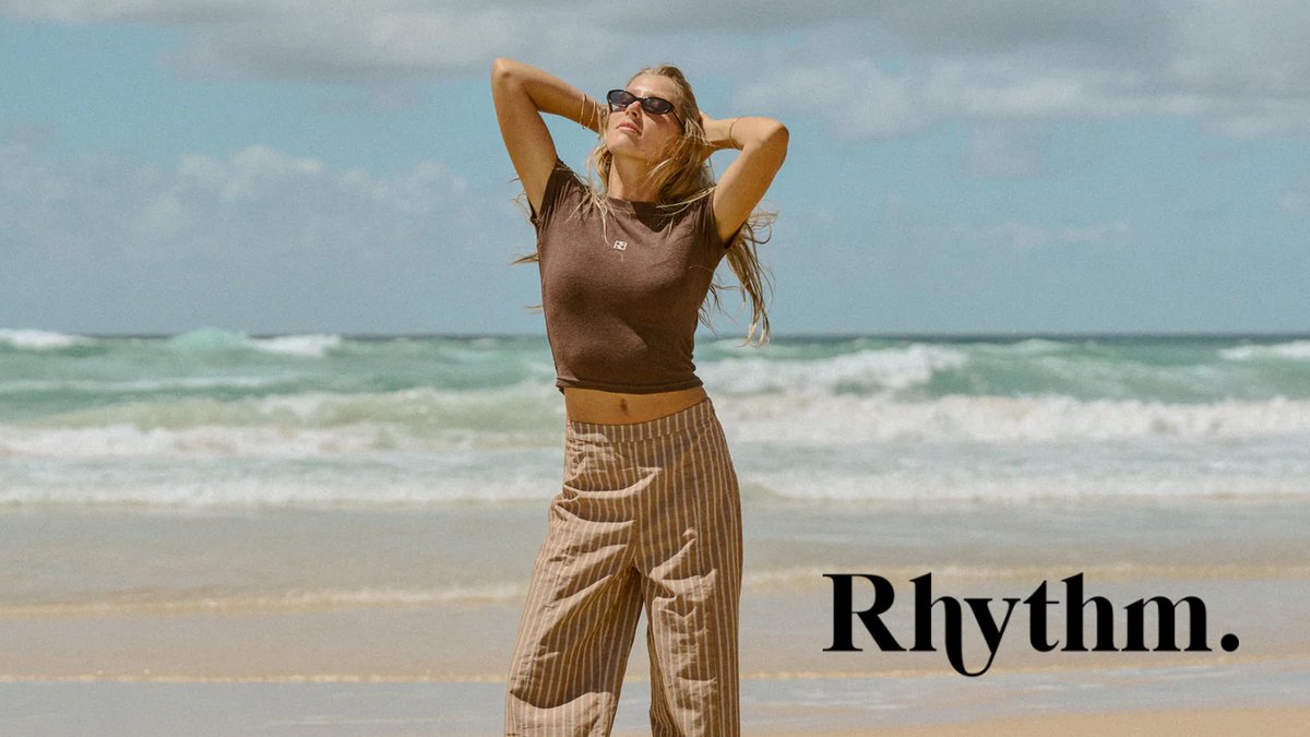Woman wearing brown crop top and beige striped pants standing on beach with ocean waves in background, modeling casual summer outdoor apparel.