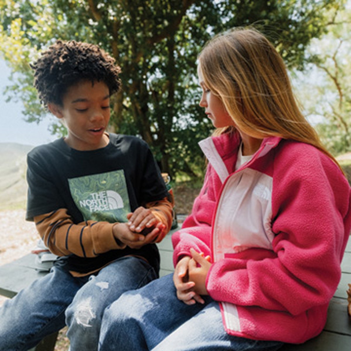 A boy and a girl sit together on an outdoor bench; the girl wears a pink fleece jacket.